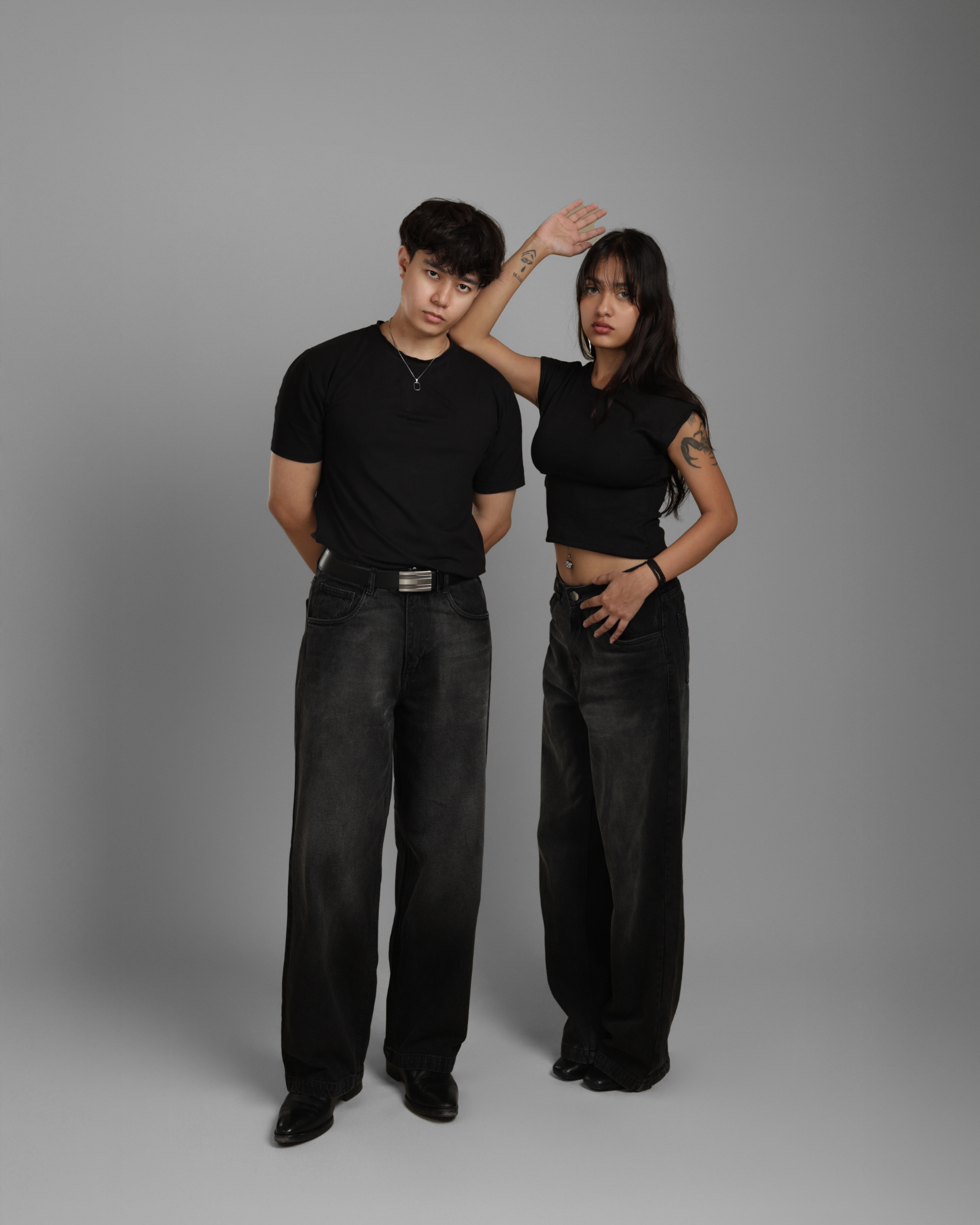 Two people wearing black outfits including black denims standing against a gray background