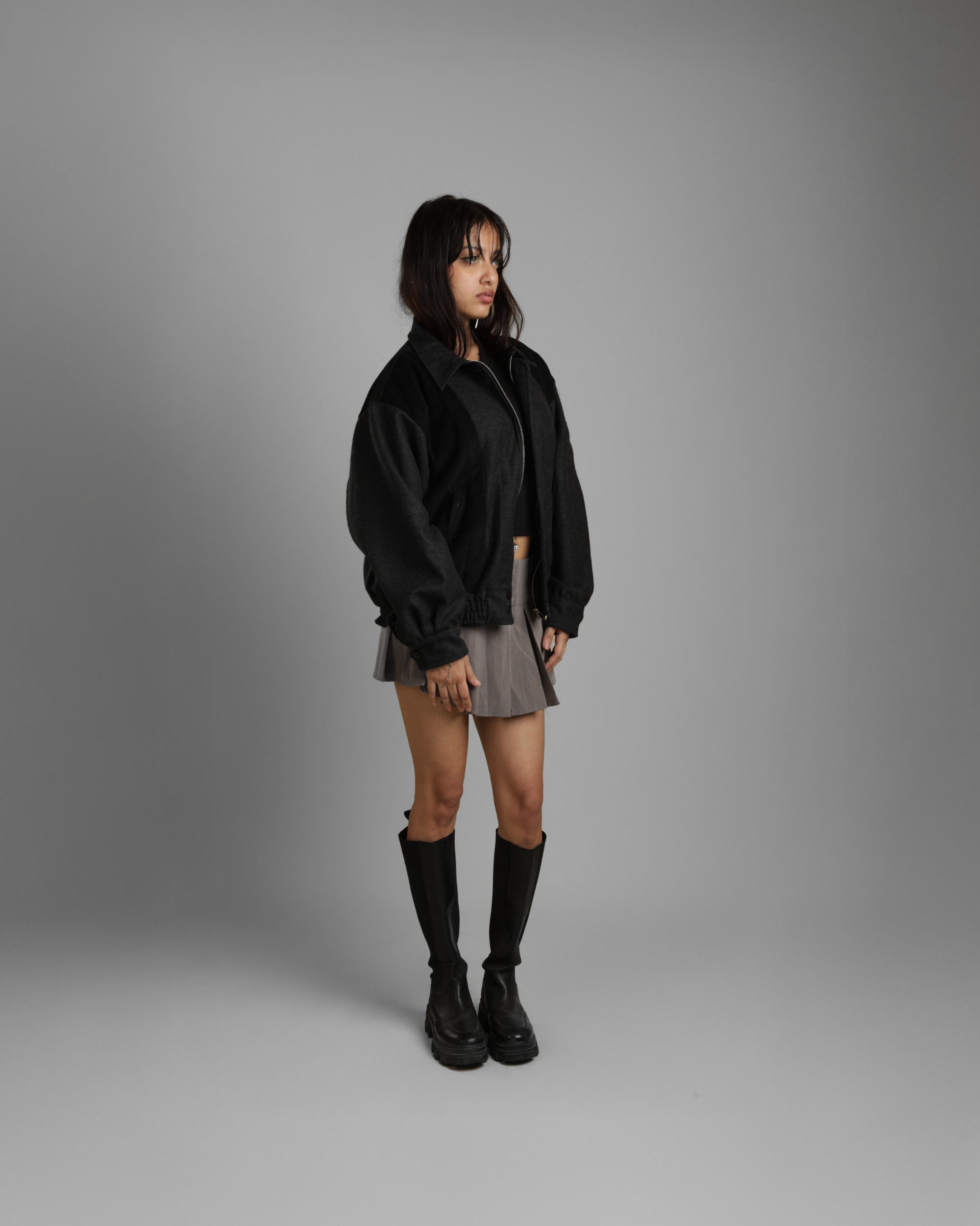 Person wearing a black zipper jacket, gray skirt, and black boots on a gray background