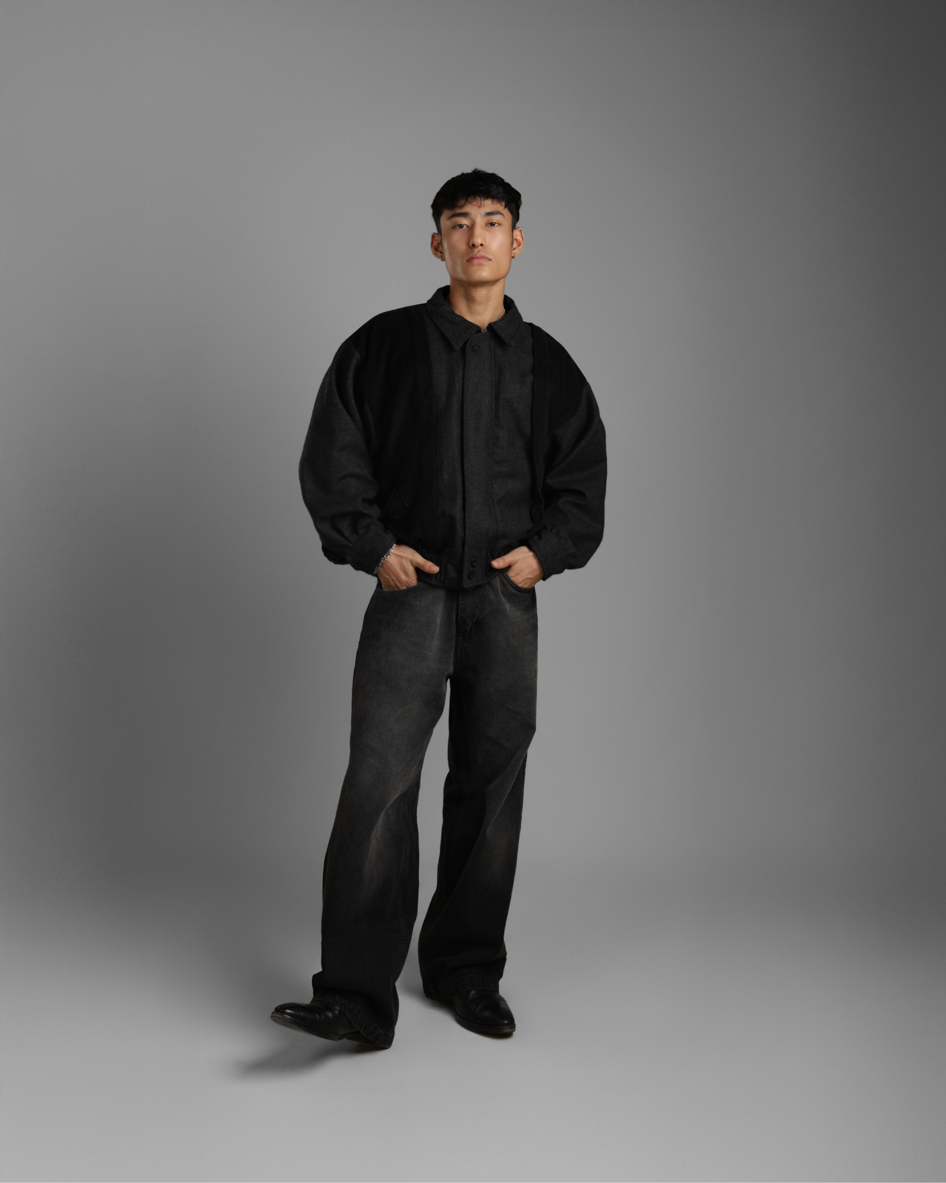 Man wearing a black grey wood corduroy zipper jacket with collar and black jeans against a gray background