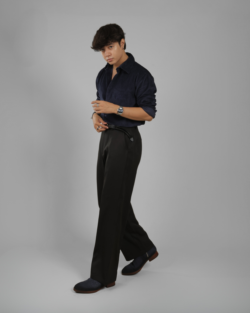 A man modeling a dark navy blue long-sleeve button-up shirt and black trousers against a solid gray studio background.