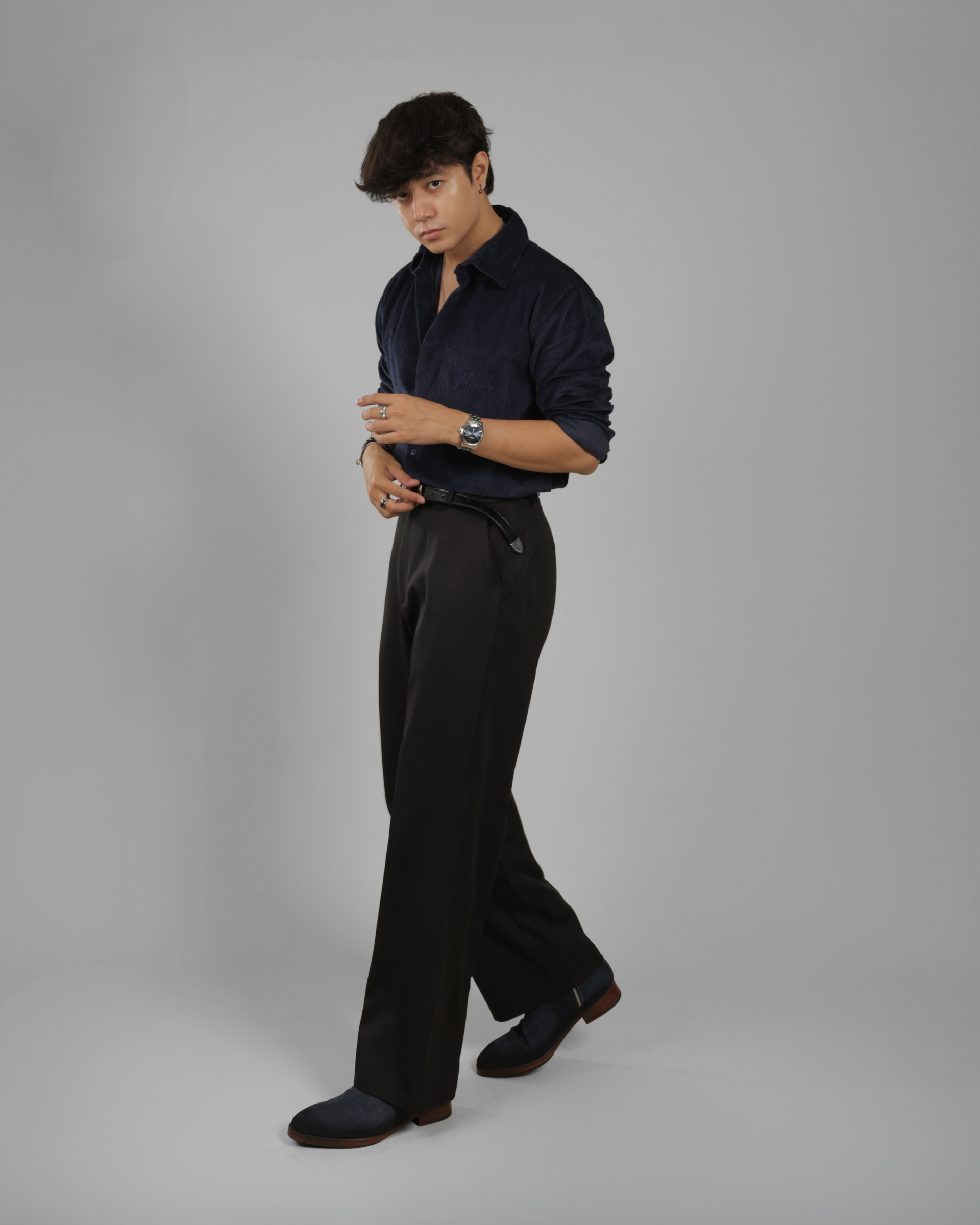 A man modeling a dark navy blue long-sleeve button-up shirt and black trousers against a solid gray studio background.