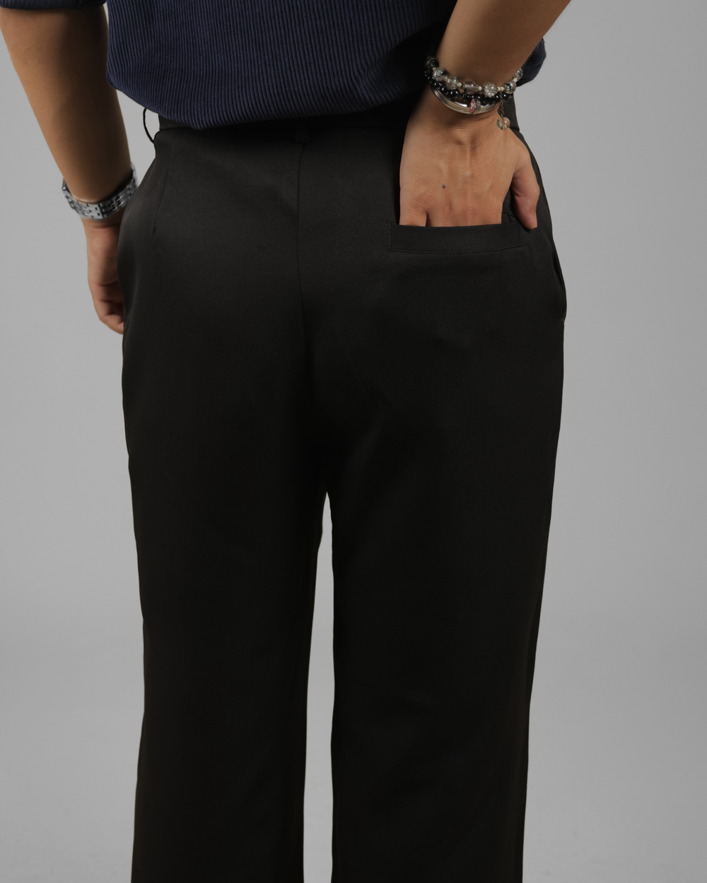A close-up shot of a person wearing black wide-leg trousers, showing the back pocket and side profile with one hand in the pocket. The person is also wearing a dark blue ribbed top and silver bracelets against a solid gray studio background.