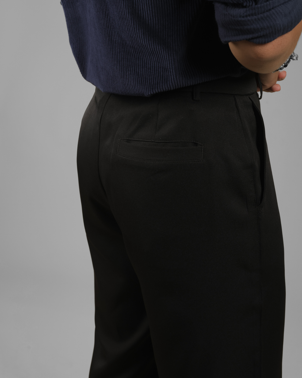 Close-up of a person wearing black trousers and a navy ribbed shirt, shown from the back against a plain gray studio background.