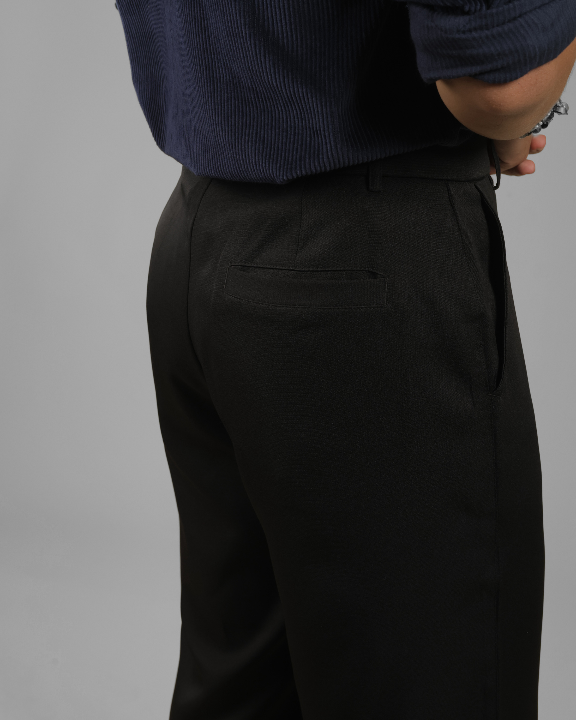 Close-up of a person wearing black trousers and a navy ribbed shirt, shown from the back against a plain gray studio background.