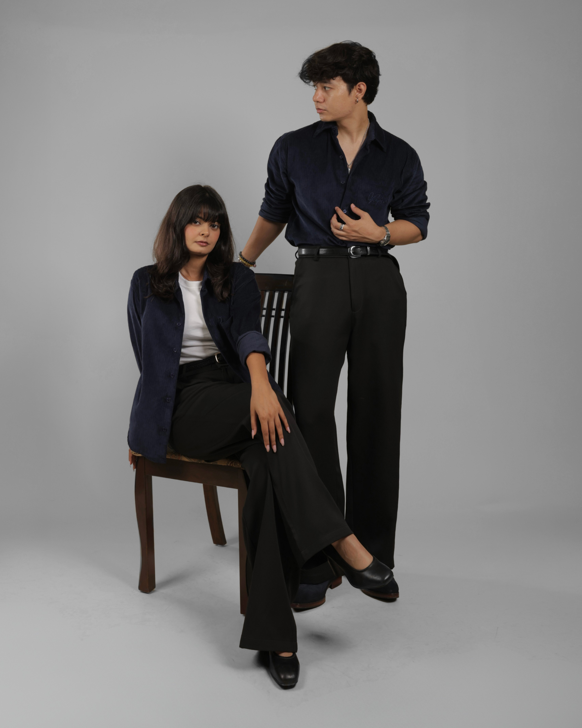 A man and a woman in dark navy and black business casual attire posing in a studio setting. The man stands while wearing a dark navy button-down shirt and black trousers, and the woman sits on a wooden chair wearing a dark navy blazer over a white top and black trousers.