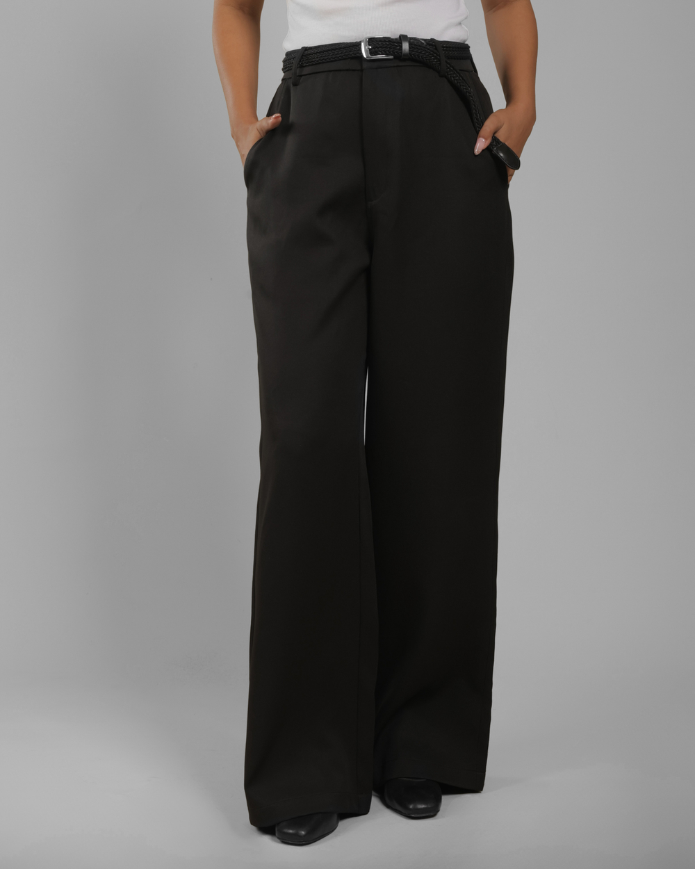 A women wearing black wide-leg trousers with a matching black belt, standing against a plain gray studio background.