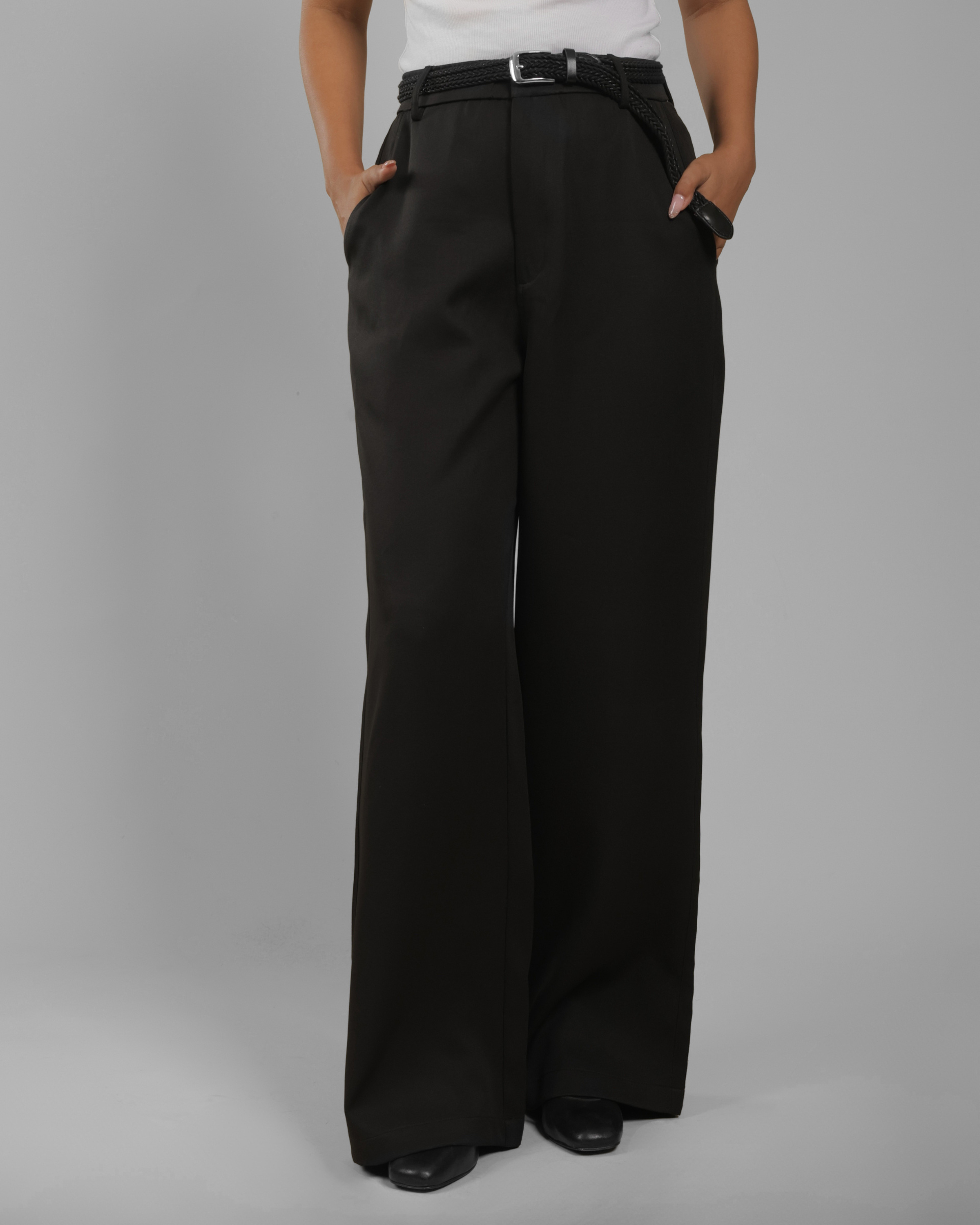 A women wearing black wide-leg trousers with a matching black belt, standing against a plain gray studio background.