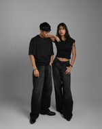 Two people wearing black outfits including black denims standing against a gray background