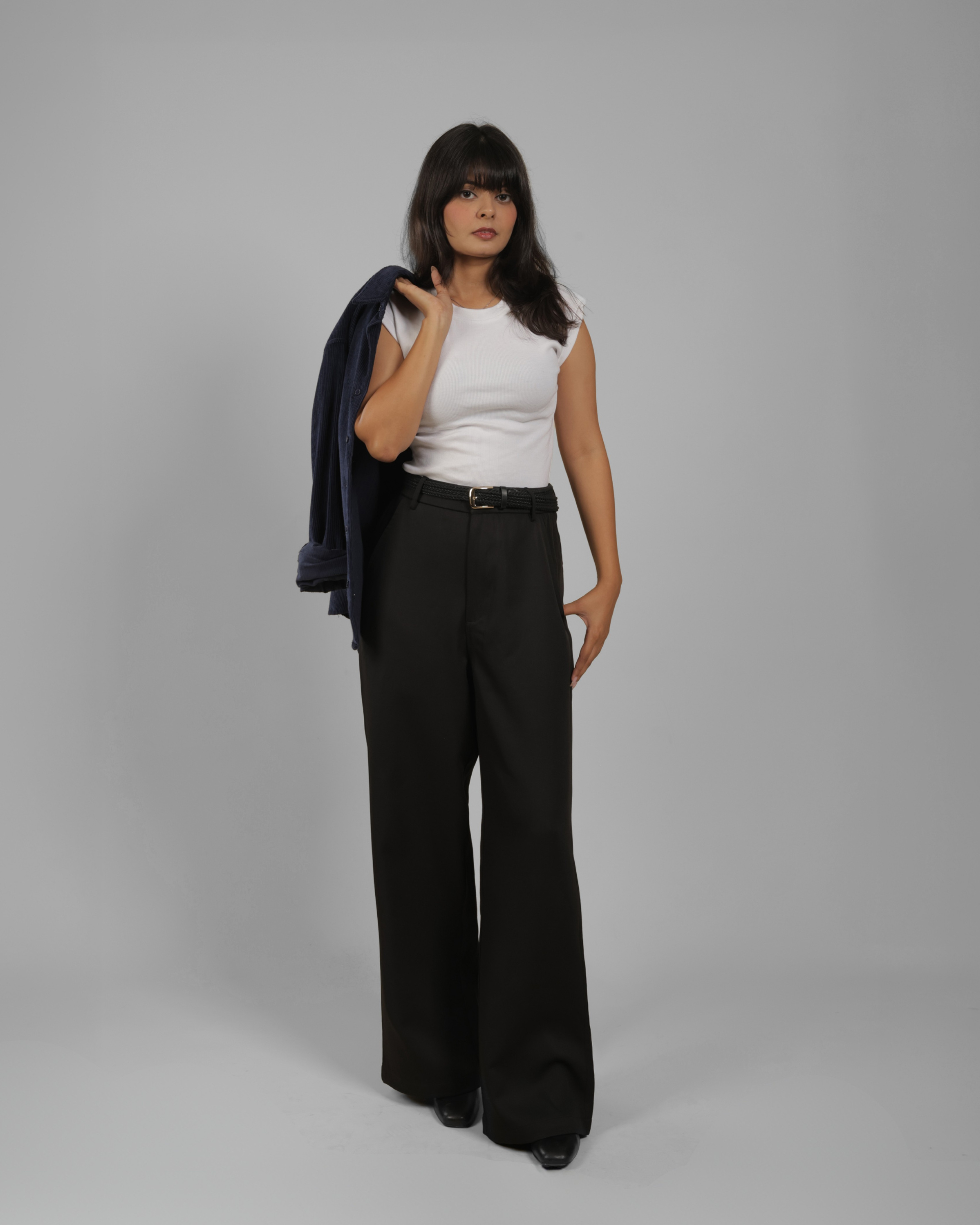 A woman stands against a light gray studio background wearing a white sleeveless top, black wide-leg trousers, and black shoes, with a dark navy shirt in her hand.