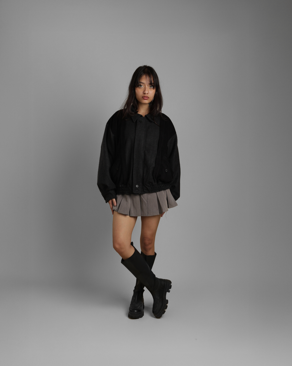 Person wearing a black jacket, gray skirt, and black boots on a gray background