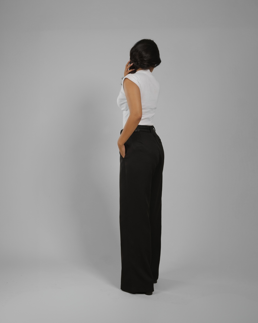 A woman shown from the back wearing a white sleeveless top and black wide-leg trousers, standing against a plain light gray studio background.