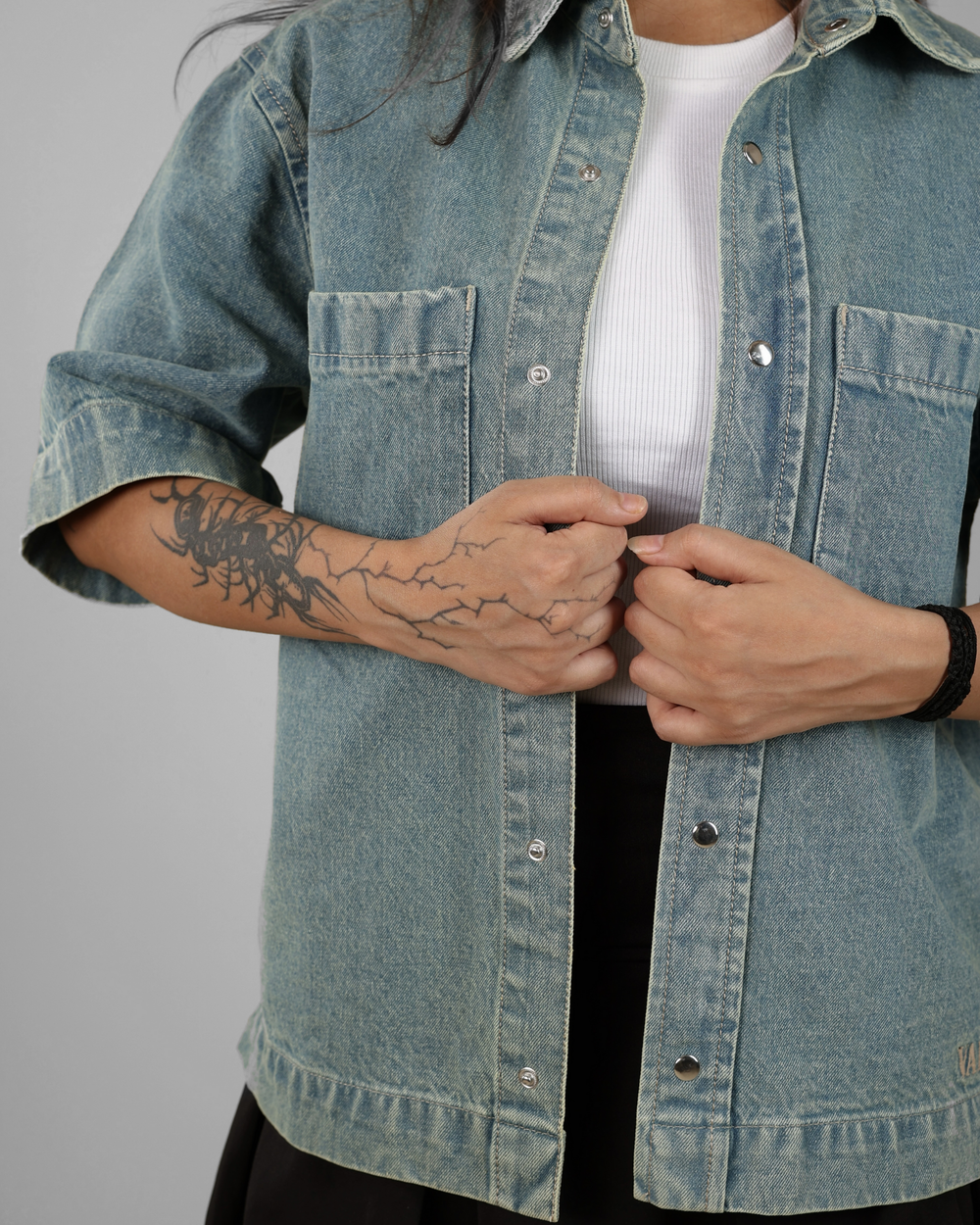 Person wearing a light blue denim jacket with a tattoo on their arm, against a plain background.