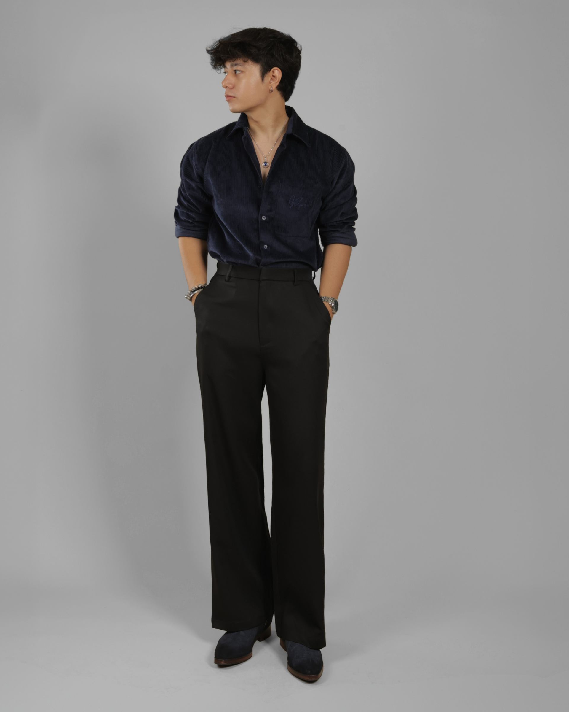 A man wearing a dark navy velvet long-sleeve button-up shirt and black wide-leg trousers, standing against a solid gray studio background.